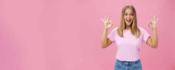 Портрет восторженной привлекательной кавказской девушки в модных футболках и джинсах, показывающих хорошо или подтверждающих жест с забавной широкой улыбкой, стоящей радостно на розовом фоне, реагируя на отличные новости - Фото, изображение