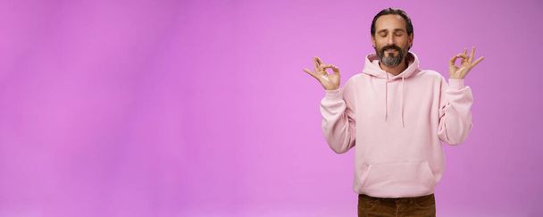 Peaceful charming hipster old man wearing cool pink hoodie close eyes breathing practice release stress meditating standing relaxed happy yoga meditation pose nirvana gesture, purple background. - Photo, Image