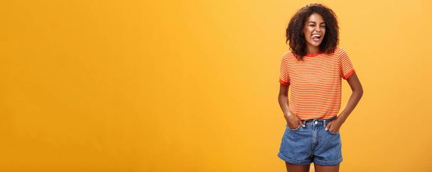 Žena se někdy chce cítit dětinsky. Radostně nadšený a charismatický mladý africký americký fena s afro účes mrkání šťastně ukazuje jazyk drží ruce v kapsách proti oranžové zdi. - Fotografie, Obrázek
