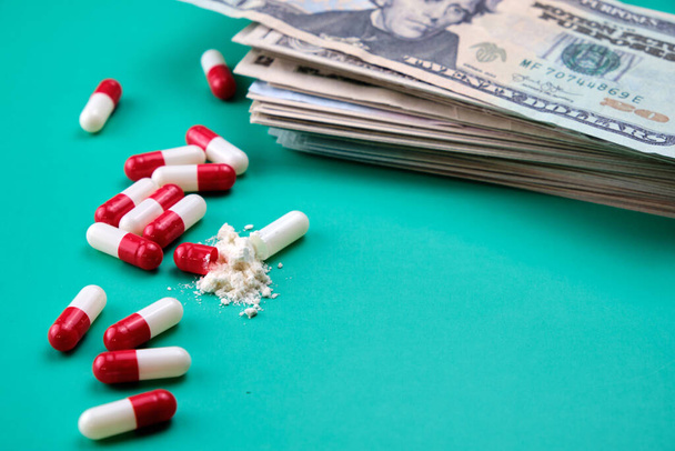 A lot of red and white capsules, one of them is open and you can see what is inside, they are on a green table with a background full of dollars. - Photo, Image