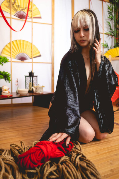 Мбаппе, стройная и молодая девушка с блондинкой в маленьком и сексуальном черном кимоно и трусиках рядом с фасонами для шибари в традиционной комнате - Фото, изображение