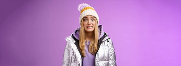 Vreemd bezorgd schattig timide blond meisje in zilveren jas hoodie winter hoed knijpen tanden popping ogen camera oeps maken fout staan nerveus iemand opvallen, paarse achtergrond. Kopieerruimte - Foto, afbeelding