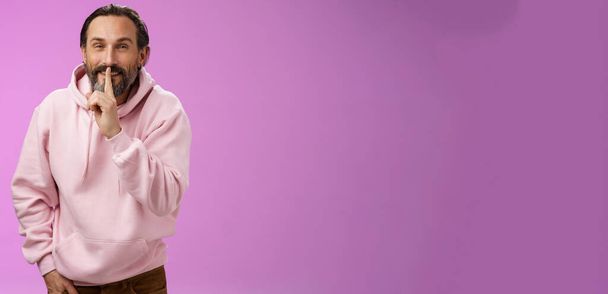 Labbra segrete. Ritratto seducente sfacciato elegante adulto barbuto ragazzo in felpa rosa con cappuccio chiedendo mantenere silenzioso spettacolo shush shh gesto indice dito premuto bocca sorridente subdolo faccia compiaciuta, avere idea. - Foto, immagini