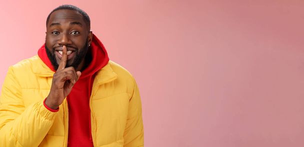 Lifestyle. Charmant jong Afrikaans-Amerikaans bebaarde man in geel jasje rood hoodie bereiden heerlijke verjaardag verrassing glimlachende show shh gebaar index vinger op mond belofte geheim te houden. - Foto, afbeelding