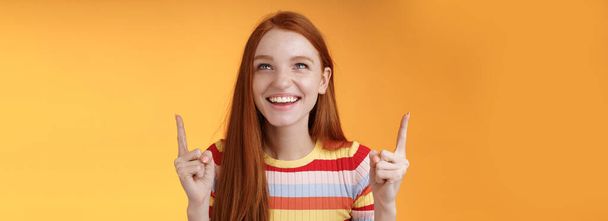 Счастливая веселая рыжая девушка веселится парк развлечений смеется радостно указывая вверх указательные пальцы смотреть вверх наслаждаться развлечением стоя оранжевый фон забавно улыбается радостно. - Фото, изображение
