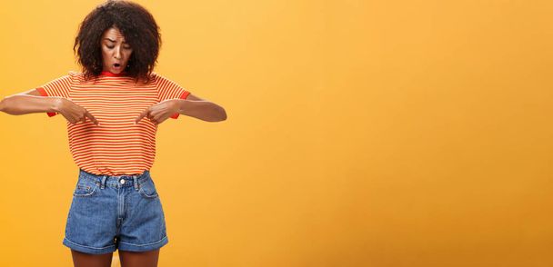 Retrato de mujer afroamericana preocupada cuestionada y sorprendida con el peinado rizado mirando y señalando el estómago o el vientre sensación de malestar y problemas con la salud sobre fondo naranja - Foto, imagen