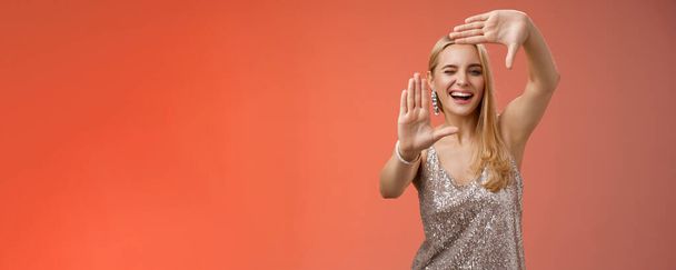 Нахальный красивый креативный европейский блондинка в серебристом блестящем платье подмигивая нахальный улыбаясь уверенный поиск вдохновение вокруг сделать рамку руки поиска место взять прохладный снимок, красный фон. - Фото, изображение