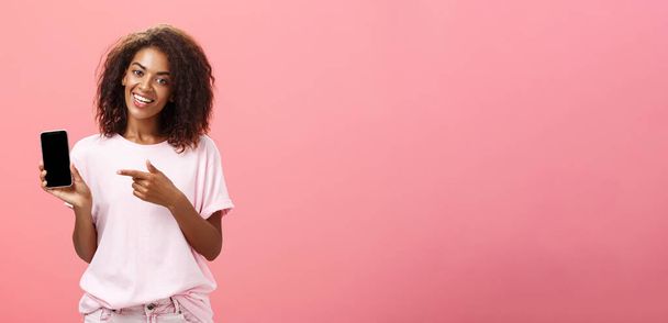 Sugira este modelo de telefone. Feliz encantador afro-americano encaracolado mulher em roupa elegante segurando smarpthone e apontando para a tela do dispositivo ao recomendar aplicativo legal sobre a parede rosa. - Foto, Imagem