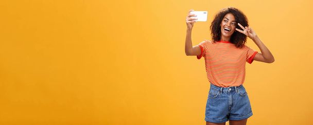 Chica haciendo video vlog con la nueva publicación de teléfonos inteligentes en Internet tratando de convertirse en famoso de pie sobre fondo naranja posando para la mirada selfie en la pantalla del gadget que muestra la paz o el gesto de victoria - Foto, imagen