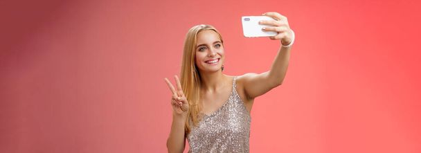 Ελκυστική θηλυκό προσφορά νεαρό ξανθό κορίτσι 25 ετών σε ασημί κομψό φόρεμα λαμβάνοντας selfie επεκτείνει βραχίονα μέχρι δείχνουν την ειρήνη χειρονομία smartphone οθόνη ρεκόρ βίντεο χαιρετισμό των οπαδών του Διαδικτύου, στέκεται κόκκινο φόντο. - Φωτογραφία, εικόνα