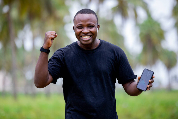Fröhlich lächelnder schwarzafrikanischer junger Mann in einem Dorf in Afrika zeigt mit dem Finger auf ein leeres Smartphone. - Foto, Bild