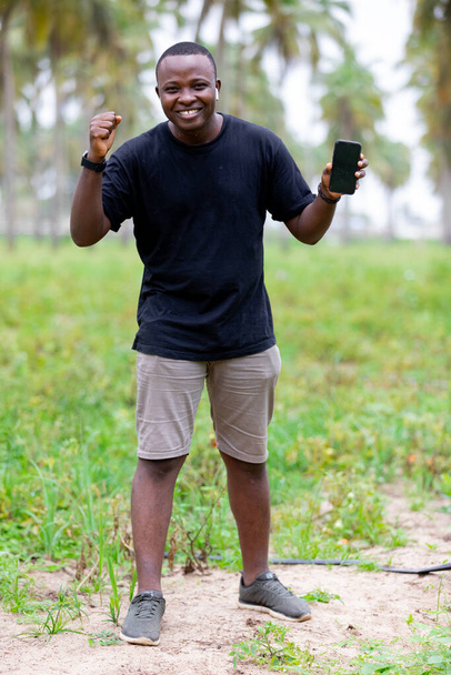 Alegre sorridente negro africano jovem na aldeia na África apontando o dedo para o smartphone tela em branco. - Foto, Imagem