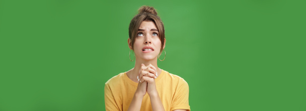 Intense nervous and faithful good-looking young woman clenching hands together over chest looking up and biting lower lip while praying god help, standing hopeful over green background. Body language - Photo, Image