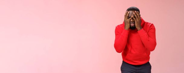 Šokovaný rozrušený mladý africký vousatý chlap v červené mikině facepalm kůže tvář ruce vykukující přes prsty smutný zničený ztratit naději, stojící smutné růžové pozadí smutek. - Fotografie, Obrázek