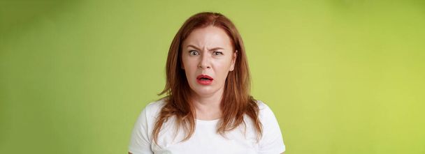 Confused shocked gasping middle-aged redhead woman cringe frustrated puzzled open mouth speechless freak out strange shocking scene stand green background perplexed disappointed. Copy space - Photo, Image