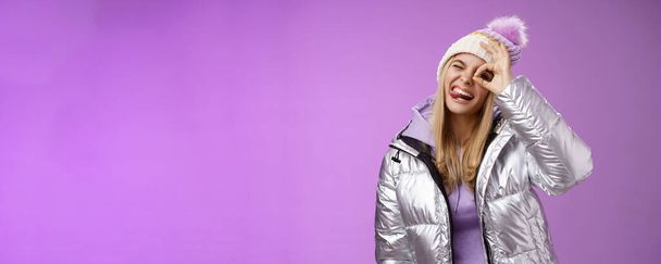 Дружня радісна біла дівчина в капелюсі тепла стильна блискуча срібна куртка нахилена голова щасливо показує язик, що посміхається, широко насолоджуючись чудовим гірськолижним курортом, подорожуючи зимою, фіолетовим фоном
. - Фото, зображення