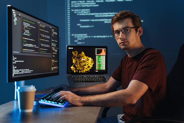 Retrato del programador masculino inteligente que trabaja en la escritura de la PC el nuevo código en su oficina casera, mirando la cámara en el fondo con la pared digital con la página de la información de la aplicación. Concepto de ciencia de datos - Foto, imagen