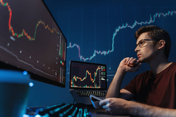 Серьезный парень криптотрейдер смотрит на экран с диаграммой подсвечника, держит ручку и блокнот, делает заметки, анализирует риски торговли криптовалютой в условиях мирового финансового кризиса - Фото, изображение