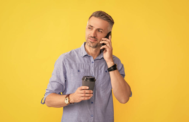 usmívající se dospělý muž mluví po telefonu s šálkem kávy na žlutém pozadí, ráno. - Fotografie, Obrázek