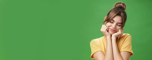 Menina europeia tímida sensual macia em camiseta amarela inclinando a cabeça no ombro tocando bochechas com as mãos fechando os olhos e sorrindo com sorriso suave sentindo nostálgico e romântico sobre fundo verde - Foto, Imagem
