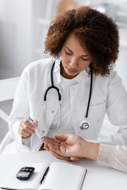 göndör afro-amerikai orvos vesz vérmintát érett beteg lándzsa toll közelében glükóz mérő az asztalon  - Fotó, kép