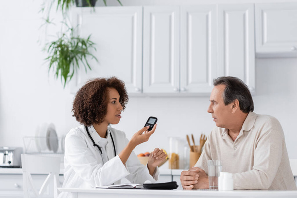 side view of curly african american doctor holding glucometer device and talking with middle aged patient - Foto, Imagen