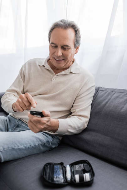 alegre hombre de mediana edad con diabetes apuntando al dispositivo medidor de glucosa y sentado en el sofá  - Foto, Imagen