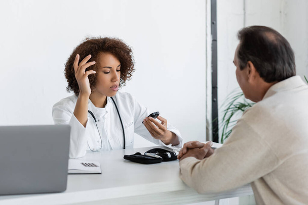 médecin afro-américain regardant le glucomètre et parlant avec un patient d'âge moyen atteint de diabète - Photo, image