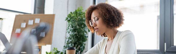 African american businesswoman sitting near blurred laptop and coffee to go in office, banner  - Фото, изображение