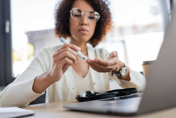 Donna d'affari africana americana offuscata che utilizza la penna lancet vicino al computer portatile e al kit per il diabete in ufficio  - Foto, immagini