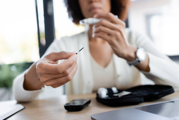 Vue recadrée d'une femme afro-américaine floue tenant une bande de test de glucomètre au bureau  - Photo, image