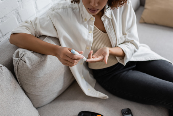 自宅でソファの上にランセットペンを使用して糖尿病を持つアフリカ系アメリカ人女性の作物ビュー  - 写真・画像