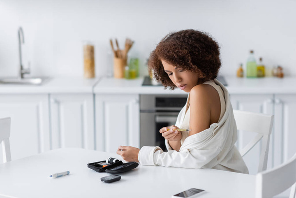 Junge afrikanisch-amerikanische Frau mit Diabetes macht Insulinspritzen in der Nähe von medizinischem Set und Smartphone in der Küche  - Foto, Bild