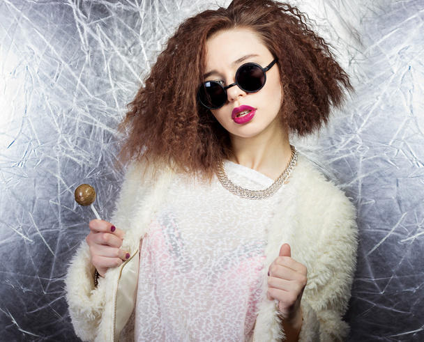 Belle fille aux cheveux bouclés et aux lèvres vives dans un manteau blanc dans les lunettes de soleil rondes avec un bonbon dans les mains, Studio shot
 - Photo, image