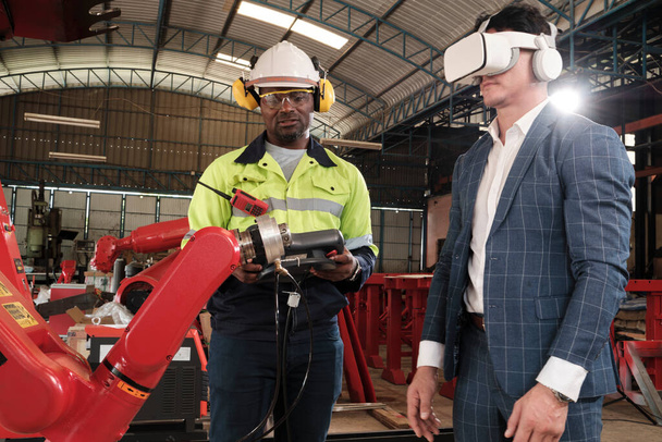 Az afro-amerikai mérnök bemutatja a robotkar vezérlését a vevőnek VR fejhallgató szimulátorral a mechanikus gyártóüzemben, intelligens ipari gépekkel és futurisztikus technológiával. - Fotó, kép