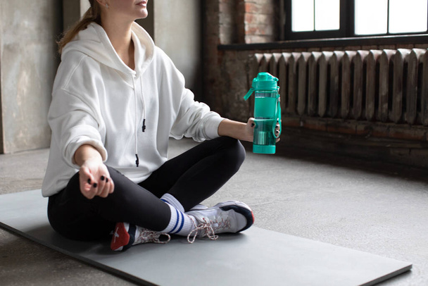The athlete is resting after training, sitting on the mat and holding a reusable sports bottle with water. Healthy lifestyle concept, reusable bottle. Copy space - Foto, imagen