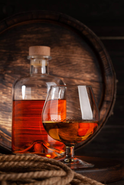 Sklenice a láhev s koňakem, whisky nebo zlatým rumem. Před starým dřevěným barelem s kopírovacím prostorem - Fotografie, Obrázek
