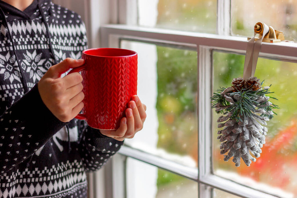 egy fiatal nő fekete pulóverben, karácsonyi mintával egy piros poharat tart a kezében az ablak mellett, ünnepi dekorációval, arc nélkül. - Fotó, kép