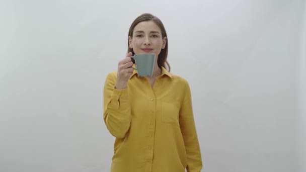 Character portrait of a young woman drinking a cup of coffee, black or green tea. Young healthy woman smelling, drinking and pointing to camera a cup of coffee or tea isolated on white background. - Footage, Video