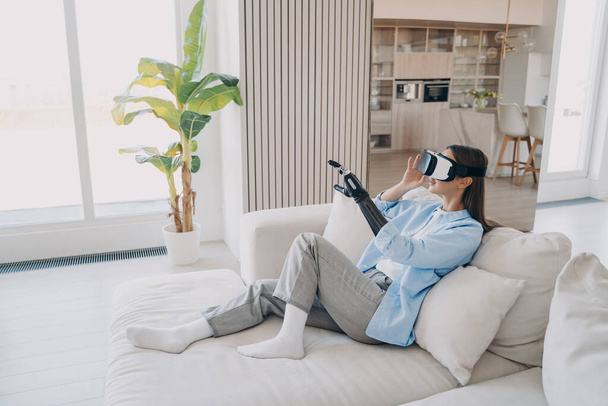Young disabled attractive european woman is sitting in living room on couch. Girl with cyber arm in vr glasses. Handicapped person gets rehabilitation at home. Excited lady touches 3d vision. - Photo, Image