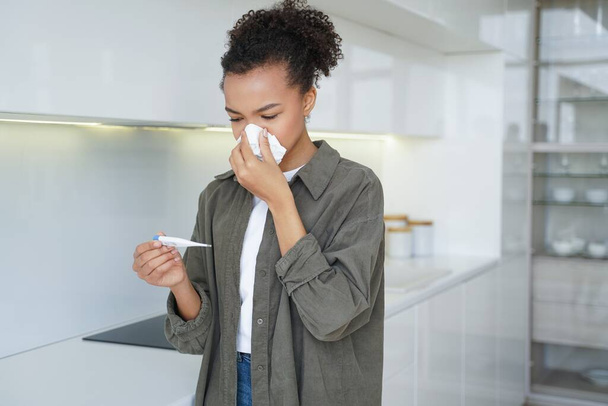 Niezdrowa młoda Afroamerykanka dmuchająca w temperaturę nosa, cierpiąca na grypę lub przeziębienie w domu. Choroba nastolatka patrzy na termometr elektroniczny, wycierając katar tkanką. - Zdjęcie, obraz