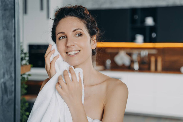 Νεαρή Ισπανίδα που σκουπίζει το πρόσωπο της με πετσέτα μετά το πλύσιμο στο σπίτι. Όμορφη χαμογελαστή γυναίκα κρατώντας πετσέτα καθαρισμού, ξήρανση του δέρματος του προσώπου με πετσέτα μετά το ντους το πρωί. Καθημερινή ρουτίνα φροντίδας του δέρματος - Φωτογραφία, εικόνα