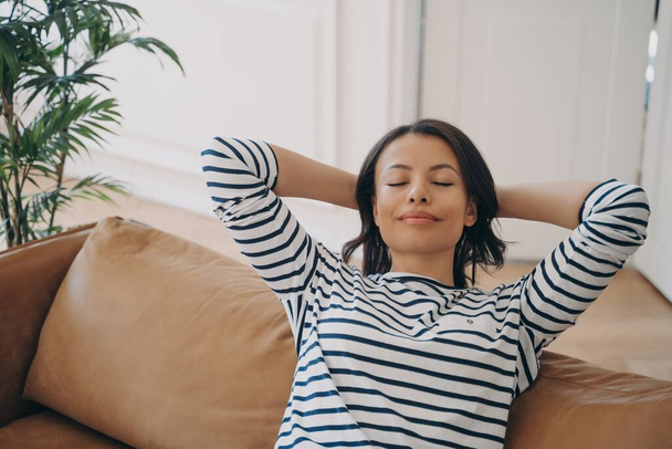 Relaxed serene woman relaxing on comfortable sofa with hands behind head at home, calm female enjoying recreation, breathing fresh air sitting on couch in living room. Stress free, wellness concept. - Photo, Image