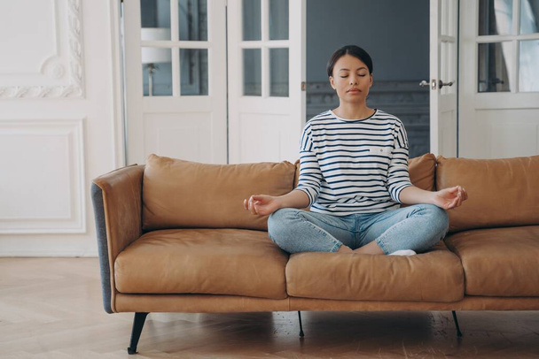 平和的なヒスパニック系の女性が自宅でヨガを練習しています。お嬢様はソファーの上で蓮の花に座ってリビングで瞑想しています。ストレス緩和とリラクゼーション。熟考と思慮深さ. - 写真・画像