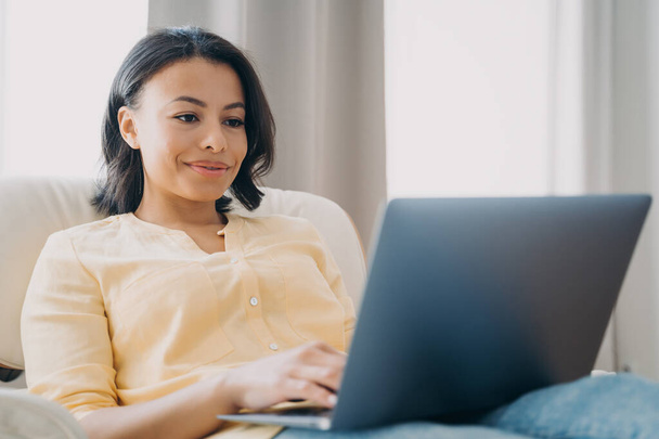 Усміхнена жінка працює на ноутбуці, сидячи в кріслі вдома, жінка дивиться на екран комп'ютера, спілкується в соціальних мережах або купує в Інтернеті. Концепція електронної комерції та електронного бізнесу
. - Фото, зображення