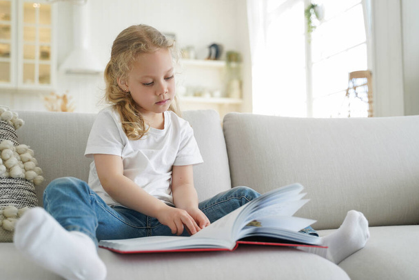 Pensive little child girl preschooler reading book, fairytale, sitting on couch in living room, small elementary kid daughter reads literature bestseller for children at home. Children's education. - Photo, Image