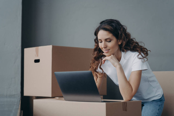 Fiatal spanyol lány kézbesítő a laptopnál dolgozik kartondobozokkal körülvéve. Üzletasszony online áruház tulajdonosa előkészíti az ügyfél megrendelések csomagok küldésére. Kisvállalkozások, e-kereskedelem. - Fotó, kép