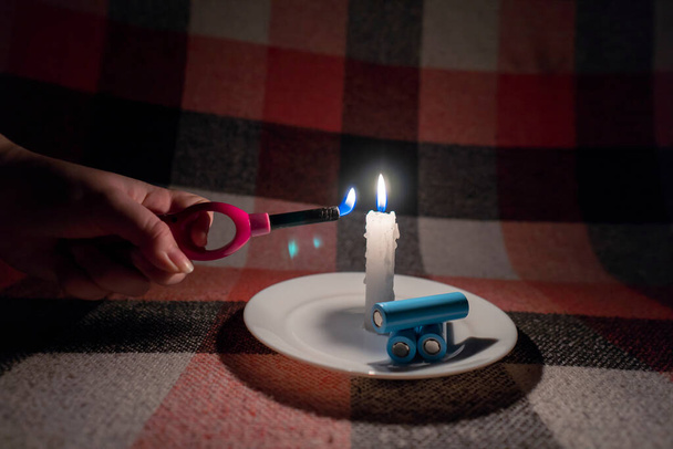 Una mujer enciende una vela con un encendedor de gas. Cerca de la vela hay tres baterías cilíndricas descargadas (primer plano). Concepto de corte de energía. Apagón. Crisis energética - Foto, imagen