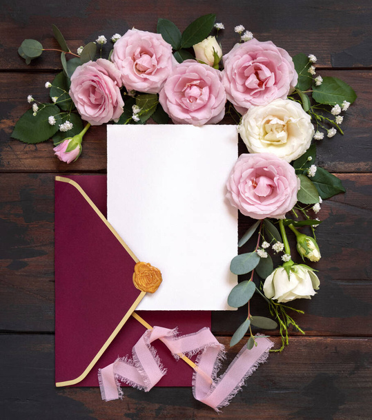 Κάρτα και κόκκινο φάκελο ανάμεσα σε ροζ και κρεμ τριαντάφυλλα σε καφέ ξύλινα πάνω όψη, γαμήλια μακέτα. Ρομαντικό σκηνικό με κάρτες, φάκελο και παστέλ λουλούδια επίπεδη θέσει. Valentines, Άνοιξη ή Ημέρα των Μητέρων έννοια - Φωτογραφία, εικόνα