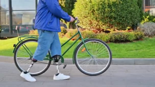 Vista de um homem com pernas protéticas. Caminhando com uma bicicleta na rua com vegetação - Filmagem, Vídeo